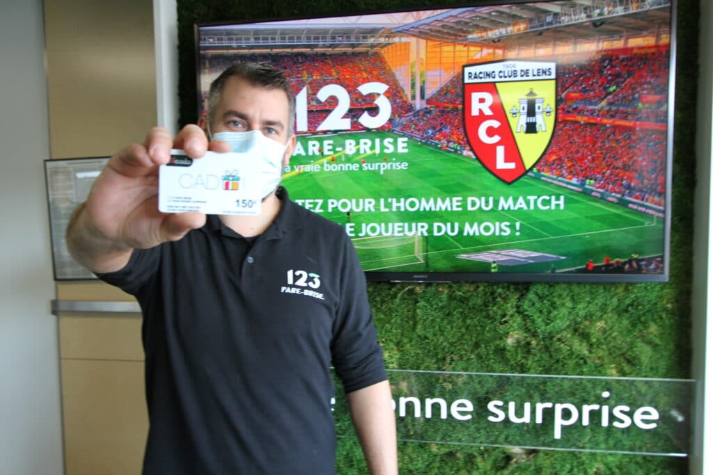 123PareBrise-Lens-joffrey-carte-cadeau Chef de centre Joffrey qui tient une carte cadeau de 150€ dans sa main devant un écran RC Lens